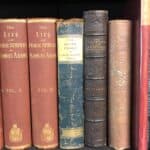 A few rare, antique books sit on a shelf covered in red, blue and leather bindings. Subjects include Samuel Adams.