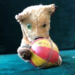 A beleagured faint brown felt-covered windup toy cat with green eyes rolls over a red and yellow tin ball.