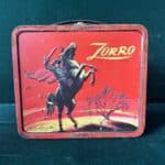 A bright red vintage lunch box depicts Zorro on his black horse Tornado, who is rising up on his back legs. Zorro raises a sword, his cape flowing in the wind behind him.