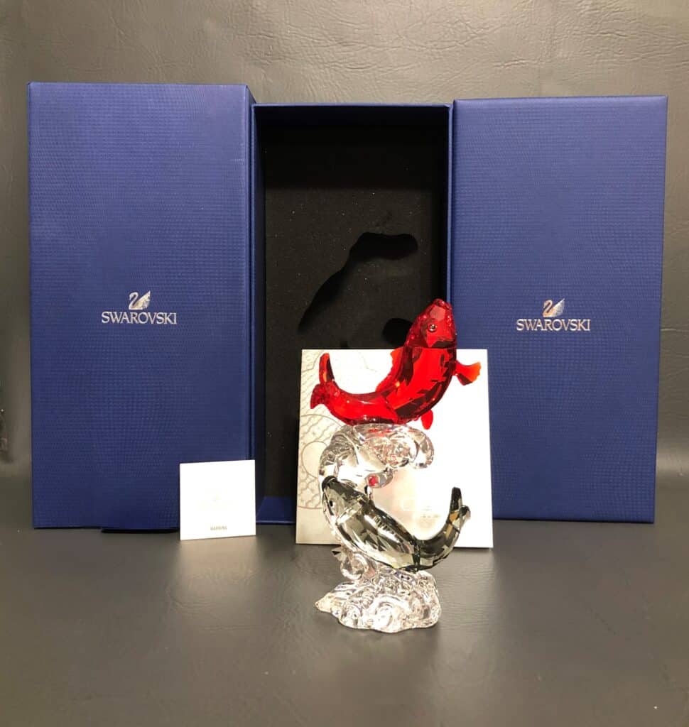 A Swarovski crystal figurine featuring a red fish twisting upward stands in front of its original blue box.