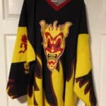 A black and yellow hockey jersey shows a yellow angry clown face with red face paint.