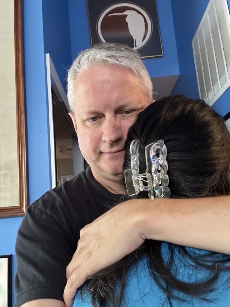 A middle-aged man with gray hair wearing a black t-shirt hugs a woman who leans into his chest.