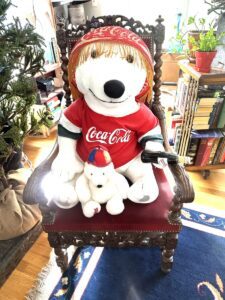 A large stuffed animal Coca-Cola bear holds a smaller one in his lap on an antique wood chair.