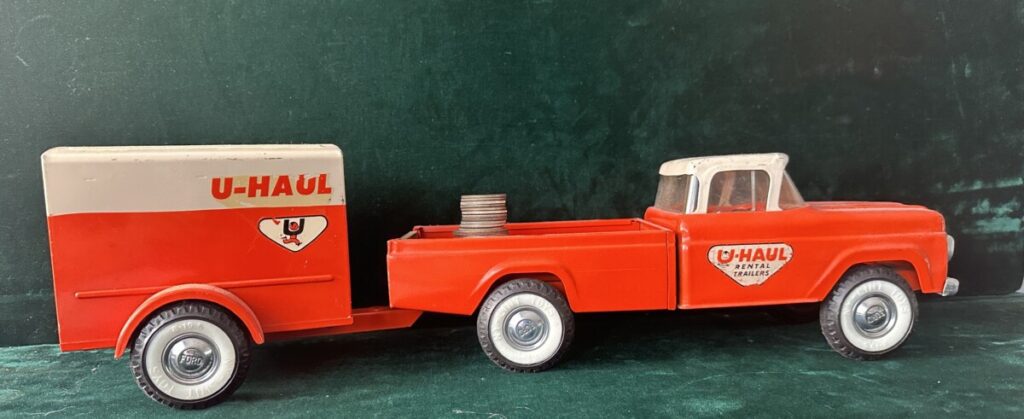 A bright orange, vintage metal U-haul toy truck and trailer roll left to right across the screen. A stack of coins sits in the flatbed.