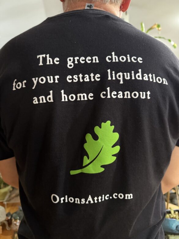 The back of a black Orion's Attic t-shirt features a green leaf and the tag line: The green choice for your estate liquidation and home cleanout