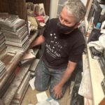 Chris Lancette of Orion's Attic kneels between hoarded bookshelves while wearing a black t-shirt. He is covered in dust.