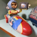 A vintage tin rocket toy with baby astronaut is marked with blue painter's tape.