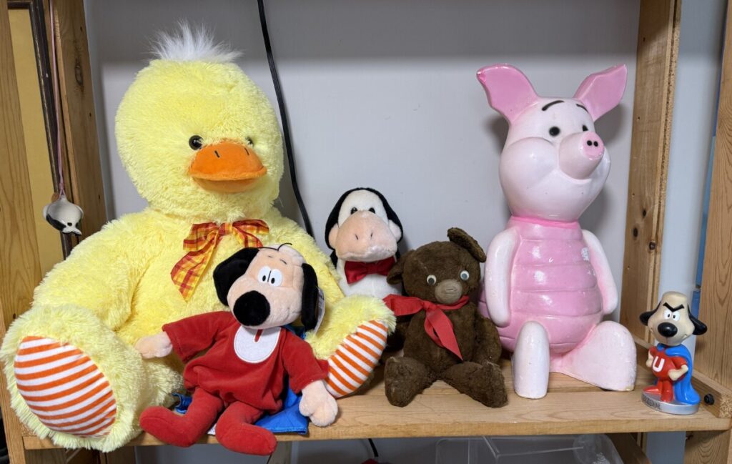 A group of stuffed animals with smiles on their faces sit on a shelf. They include a large duck, Opus, a teddy bear, and Piglet.