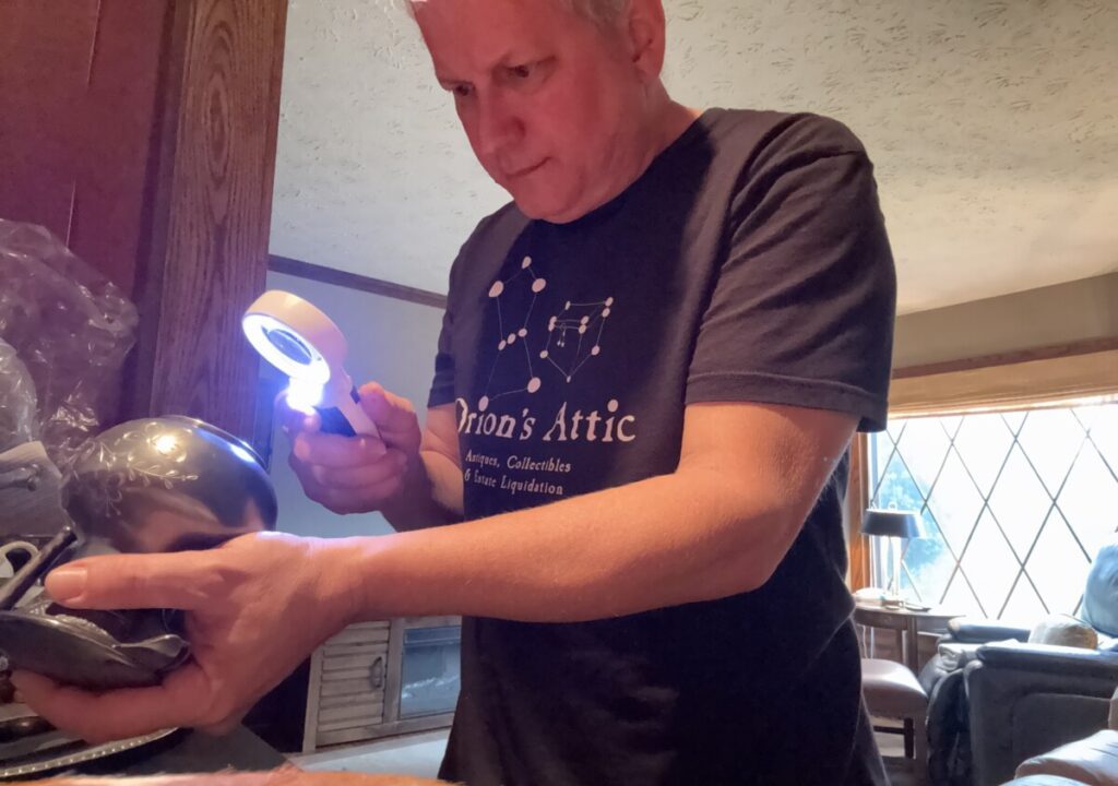Orion's Attic co-owner Chris Lancette examines a silver dish with a magnifying glass looking for sterling silver marks.